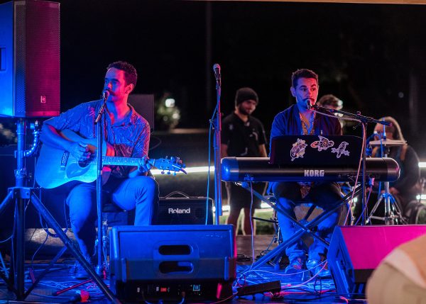 Two males sitting on stage, one playing a guitar, the other singing and playing keyboard
