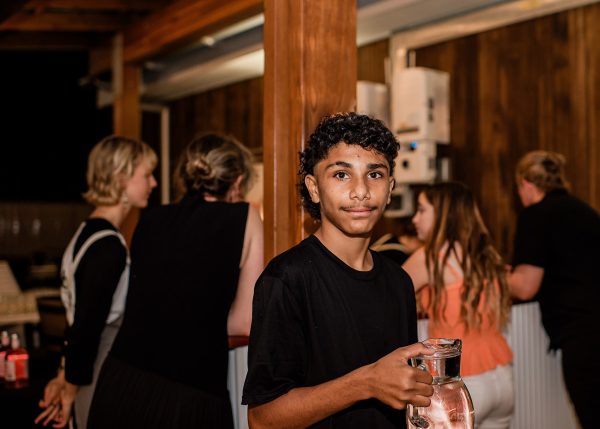 A teenager with dark hair and a black shirt carrying a water jug at a gala