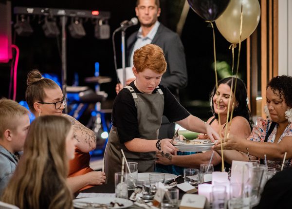 A young man with red hair picking up plates from a table of guests at the gala