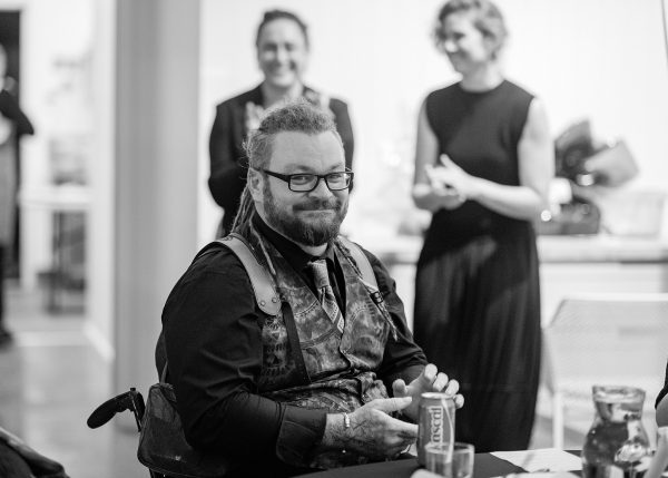 A black and white photo of a man with a beard and dreadlocks wearing a vest in a wheelchair smiling. Two females in the background smiling 