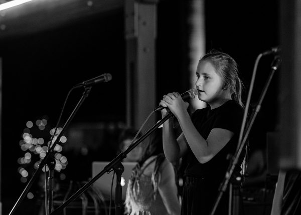 A black and white image of a young female singing into a microphone on stage, looking sidewards