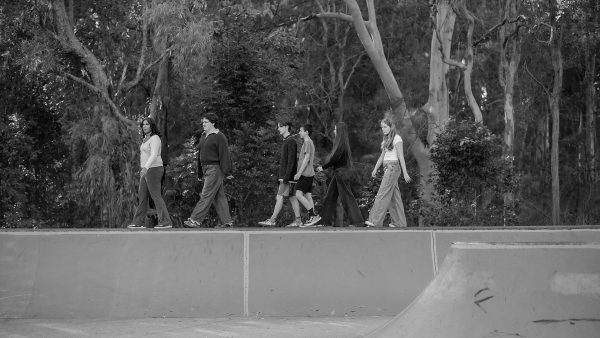 black and white image of 6 young people walking along the top of a skate ramp with trees in the background