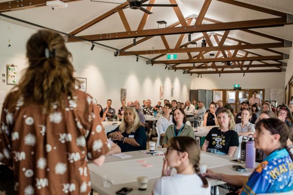 Lady in brown dress with her back to the camera speaking to a room of adults sitting at tables