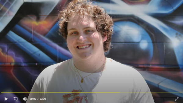Image of a young Caucasian male in a white shirt smiling in front of a grafitti wall