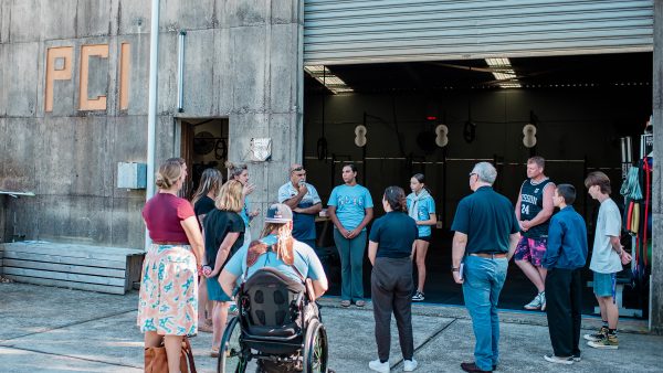 a group of people standing in a circle out the front of a shed with an open door