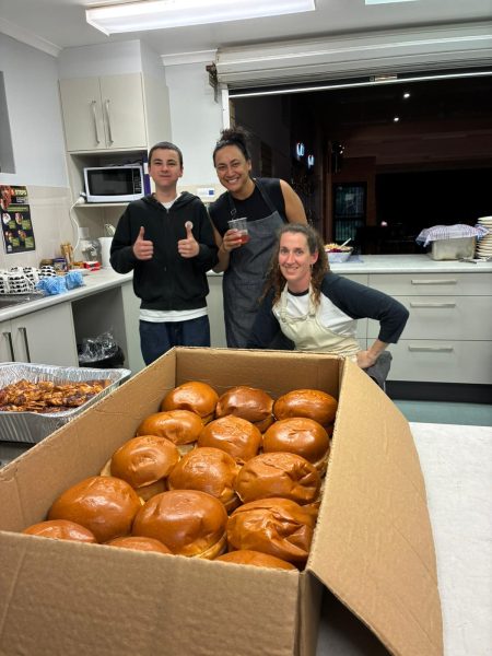 Two woman and a male teenager standing in a kitchen with thumbs up in front of a big box of breadrolls