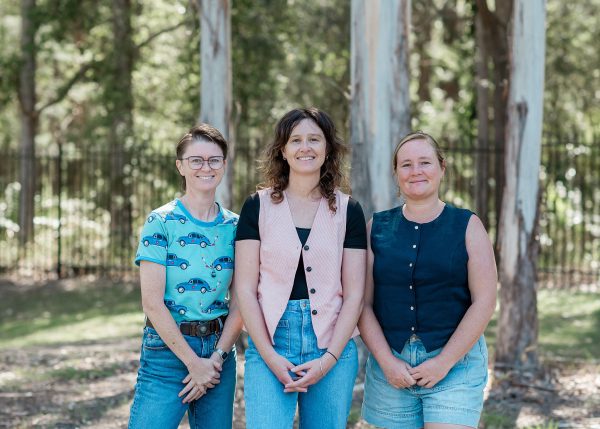 three women standing with their hands in front of them with a background of trees and grennery